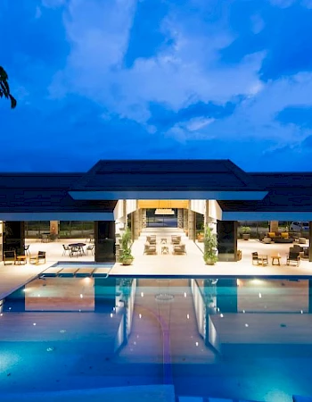A luxurious modern building at night with a large illuminated pool in front, surrounded by seating and greenery.