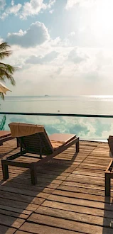 A serene poolside scene with wooden lounge chairs, umbrellas, and palm trees overlooking a calm sea under a partly cloudy sky.