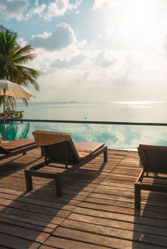 A tropical scene with lounge chairs, umbrellas, and palm trees by a pool, overlooking the ocean under a partly cloudy sky and sunlight.