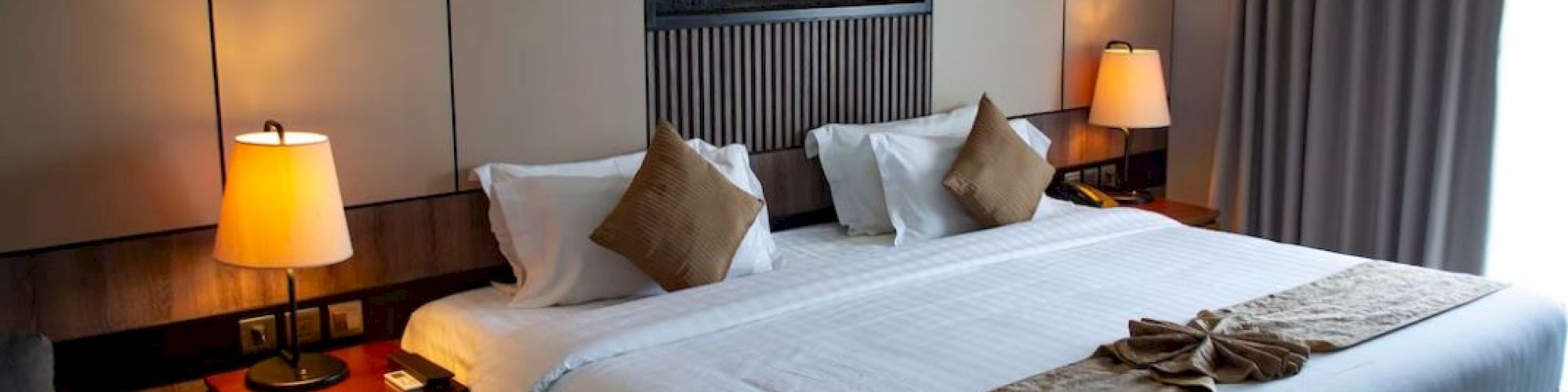 A modern hotel room with a large bed, decorative pillows, warm lighting, and a small artwork above the bed. Curtains cover a window.