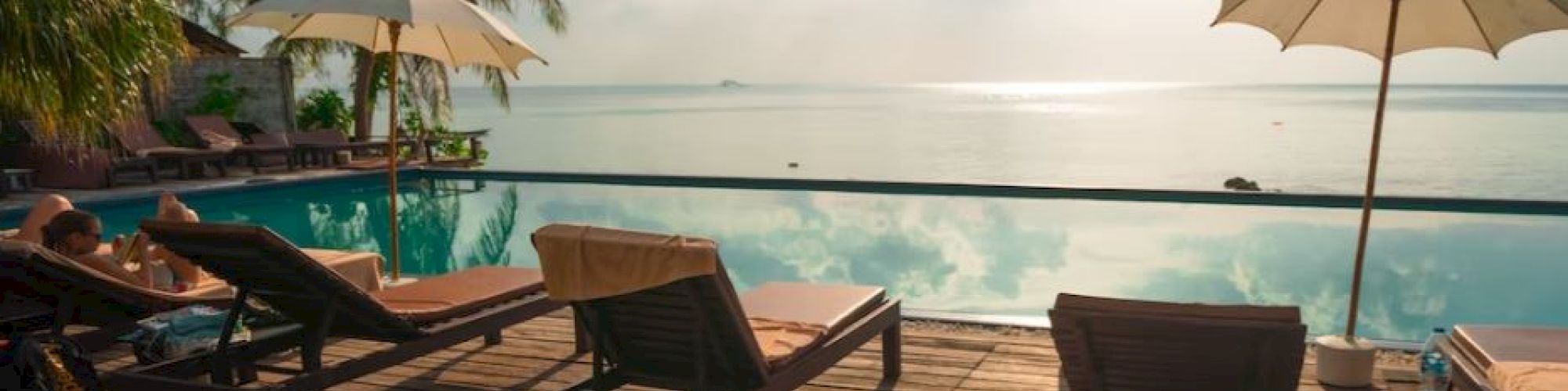 A serene poolside scene with lounge chairs, umbrellas, palm trees, and a view of the ocean under a partly cloudy sky.