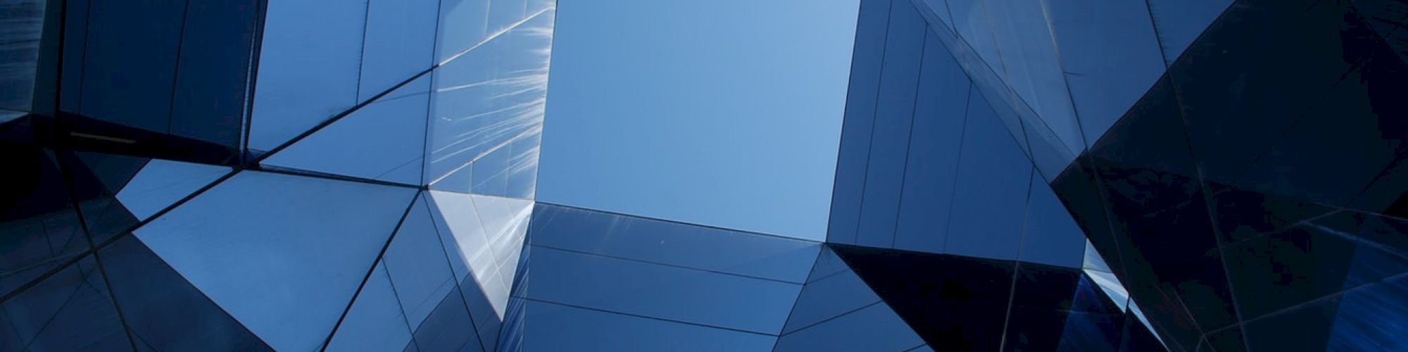 The image shows a geometric arrangement of reflective glass skyscrapers with a patch of clear blue sky visible in the center.