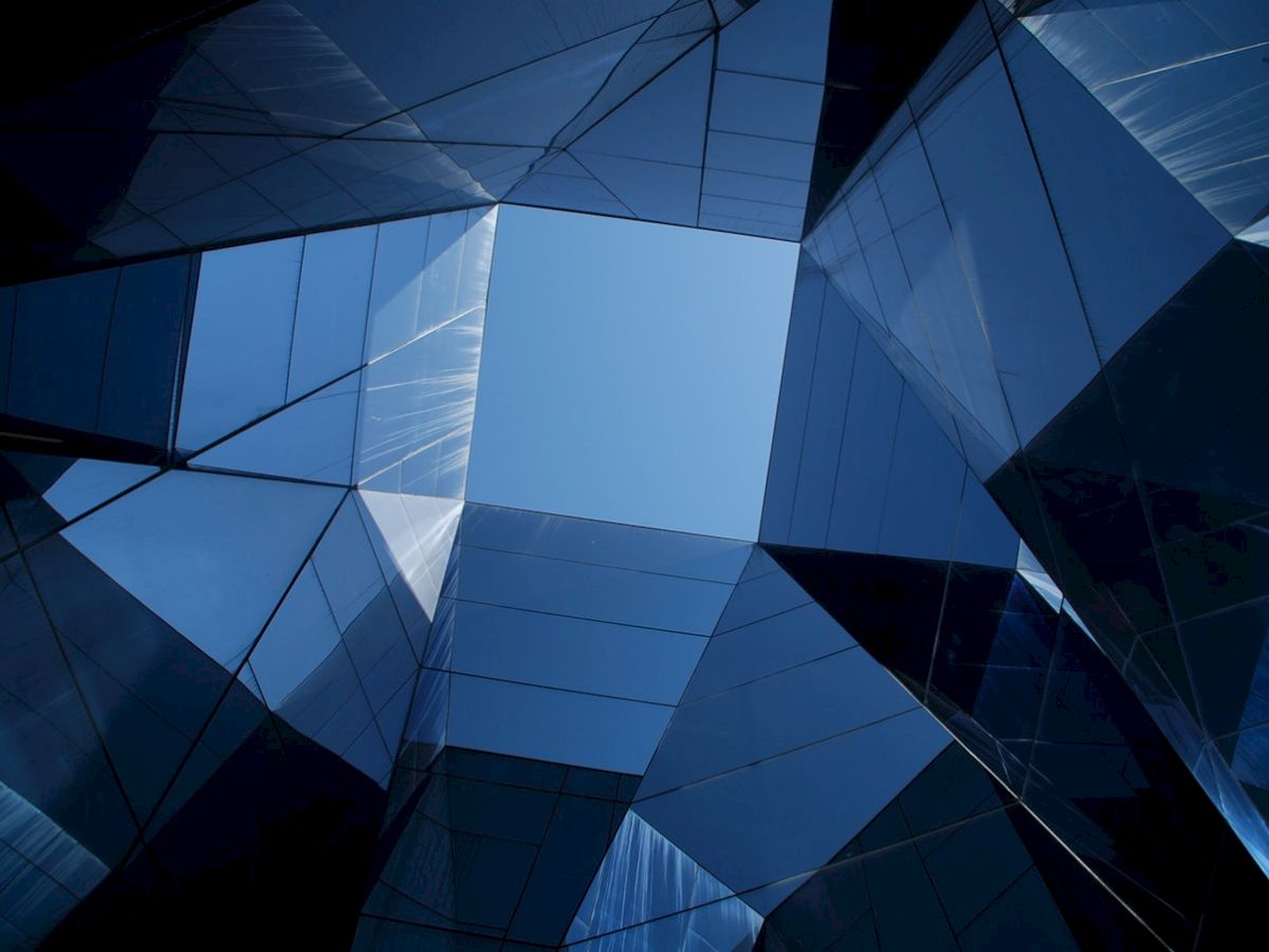 The image shows abstract architecture with reflective glass surfaces, forming geometric shapes that frame a patch of blue sky in the center.