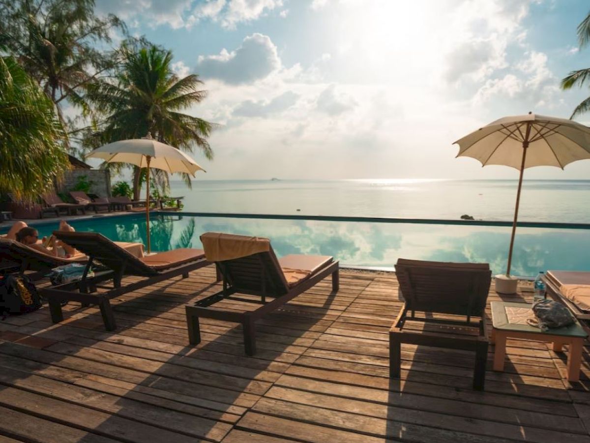 A serene poolside scene with sun loungers, umbrellas, palm trees, and an ocean view under a bright sky.
