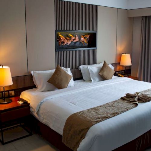 The image shows a hotel room with a large bed, decorative pillows, bedside lamps, and a wall feature. The curtains are drawn to the side.