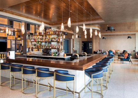 A modern bar with a well-stocked shelf, surrounded by blue bar stools and stylish lighting, in an inviting and spacious lounge area.