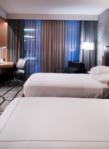 The image shows a modern hotel room with two beds, a desk and chair, wall-mounted TV, and stylish lighting and decor.