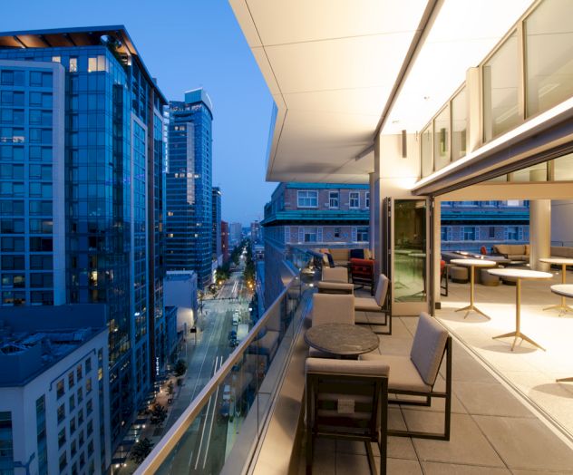 A modern urban balcony overlooks a city street, featuring glass railings, comfortable seating, and outdoor tables during the evening.