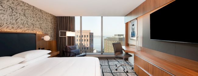 Modern hotel room with large bed, patterned wall, desk with chair, big TV, and a window showing city buildings and water.