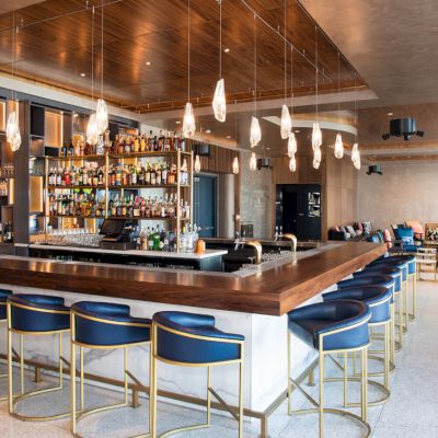 A modern bar with a wooden counter, blue cushioned stools, pendant lights, and a wide selection of bottles on display. Cozy seating area nearby.