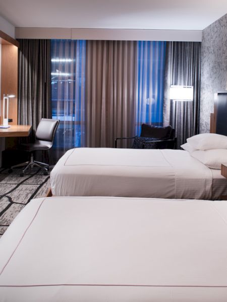 A modern hotel room with two beds, a desk, a TV, and stylish lighting. The decor is contemporary with patterned carpet and walls.