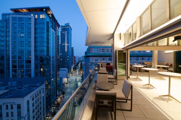 A modern cityscape at dusk is visible from a balcony with seating and tables, overlooking tall buildings along a street.