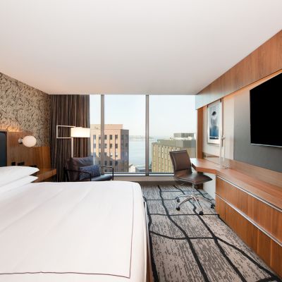 A modern hotel room with a large bed, desk, chair, flat-screen TV, and city view through a window.