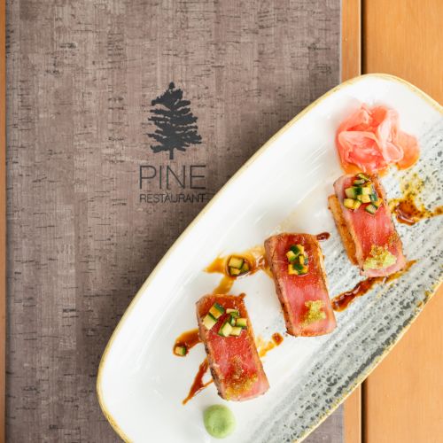 A plated dish of bacon-wrapped pieces with cucumber on sauce, next to a &ldquo;PINE Restaurant&rdquo; sign.