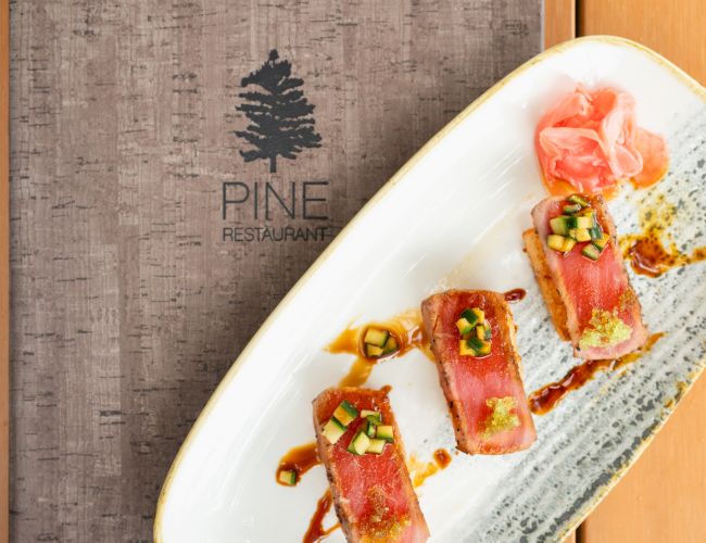 A plated dish of bacon-wrapped pieces with cucumber on sauce, next to a &ldquo;PINE Restaurant&rdquo; sign.
