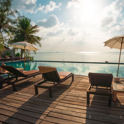 A tranquil beach setting with lounge chairs, umbrellas, and a pool overlooking the ocean under a bright sky with clouds.