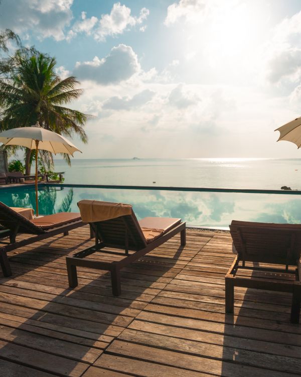 A tranquil beach setting with lounge chairs, umbrellas, and a pool overlooking the ocean under a bright sky with clouds.
