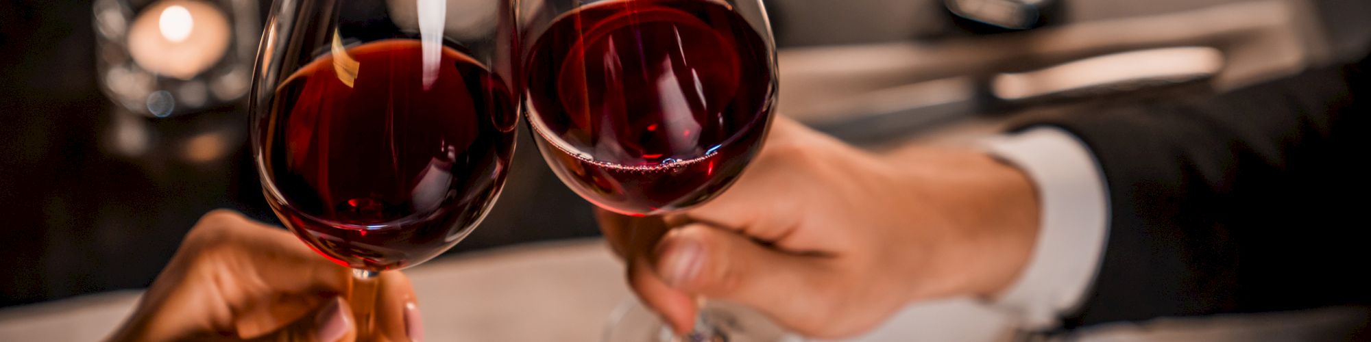 Two people clink glasses of red wine over a table set with food and lit candles, creating a cozy and romantic atmosphere.