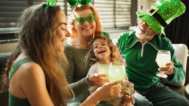 A joyful group celebrates with drinks, wearing green clothes and hats, possibly for St. Patrick's Day, smiling and enjoying together.