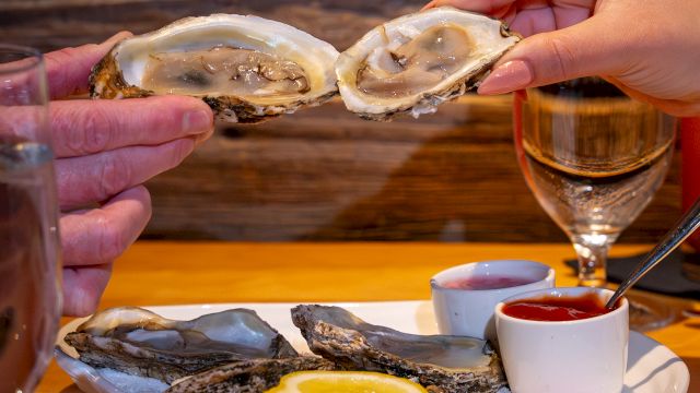 Two hands holding oyster shells above a plate with oysters, lemon, and sauces; glasses of white wine are visible.