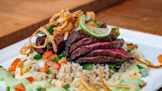 A plated dish with sliced meat, crispy onion strips, vegetables, and a grain base, garnished with sauce, on a wooden table.