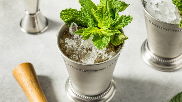 A mint julep cocktail with crushed ice and fresh mint leaves in a metal cup, sitting next to a wooden muddler on a light surface.