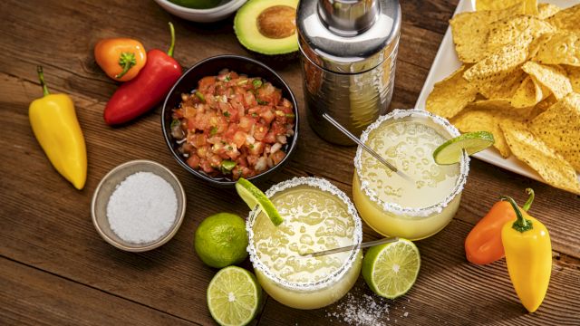 The image shows margaritas with lime, a shaker, salsa, avocados, peppers, and tortilla chips on a wooden table.