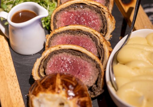 The image shows slices of beef Wellington, mashed potatoes, gravy, and greens, served on a wooden board.