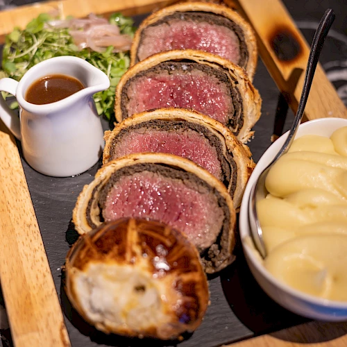 The image shows slices of beef Wellington, mashed potatoes, gravy, and greens, served on a wooden board.