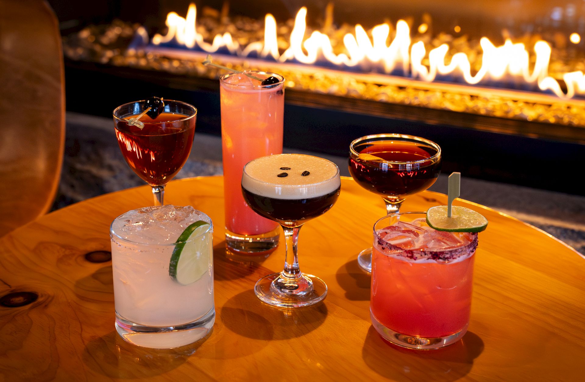 A variety of cocktails on a wooden table in front of a cozy fireplace, garnished with lime, cherries, and espresso beans.