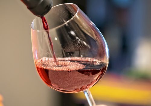 A glass is being filled with red wine from a bottle, with bubbles visible on the wine’s surface in a softly lit setting.