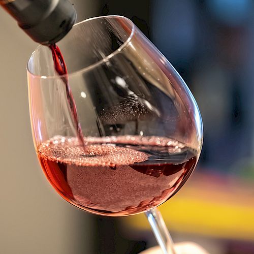 A glass is being filled with red wine from a bottle, with bubbles visible on the wine’s surface in a softly lit setting.
