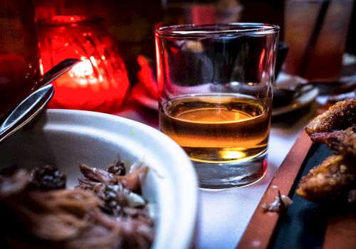 A glass of amber liquid, possibly whiskey, is on a table with a lit candle, food, and utensils, creating a cozy dining ambiance.