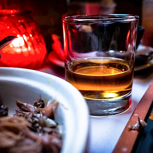 A glass of amber liquid, possibly whiskey, is on a table with a lit candle, food, and utensils, creating a cozy dining ambiance.