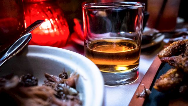 A glass of amber liquid, possibly whiskey, is on a table with a lit candle, food, and utensils, creating a cozy dining ambiance.
