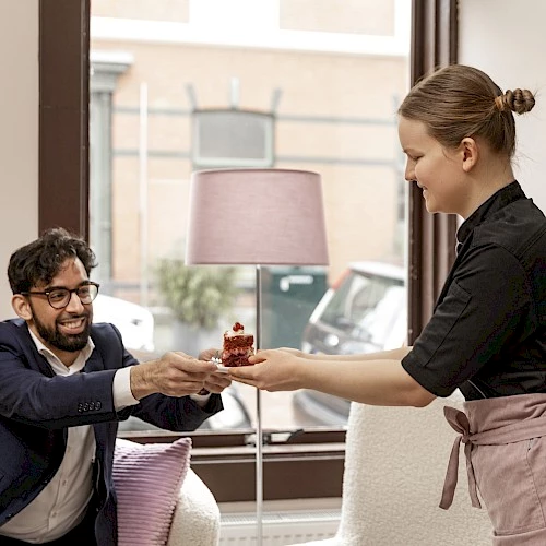 A seated man receives a dessert from a woman in a cozy room with a lamp by the window.