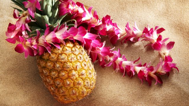 A pineapple with a pink and white flower lei draped over it, set on a textured beige surface.