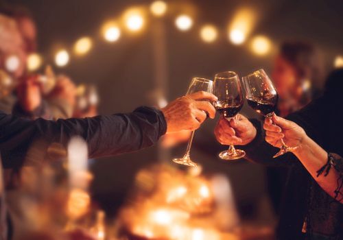 People are toasting with wine glasses at a gathering or celebration, with warm lighting and a blurred background.