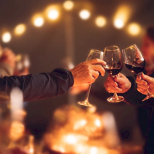 People are toasting with wine glasses at a gathering or celebration, with warm lighting and a blurred background.