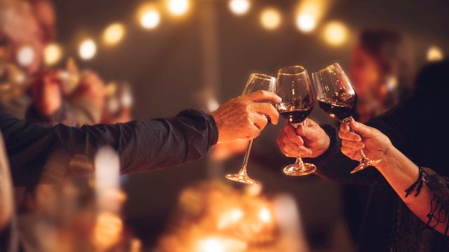 People are toasting with wine glasses at a gathering or celebration, with warm lighting and a blurred background.
