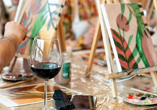People are painting on easels with abstract designs. A glass of red wine and a phone are on the table.