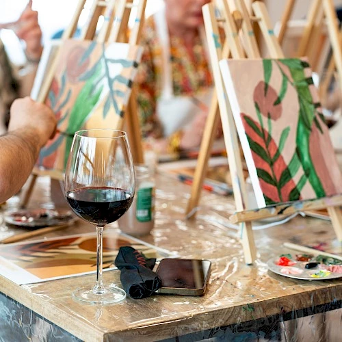 People are painting on easels with abstract designs. A glass of red wine and a phone are on the table.