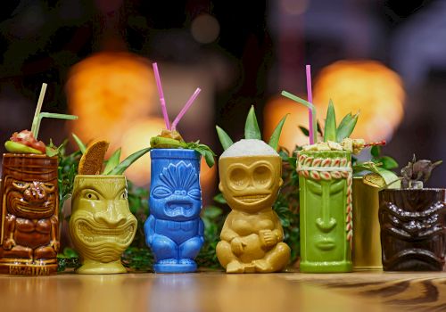 Colorful tiki mugs with straws are filled with tropical drinks, garnished with fruits and leaves, arranged on a table.