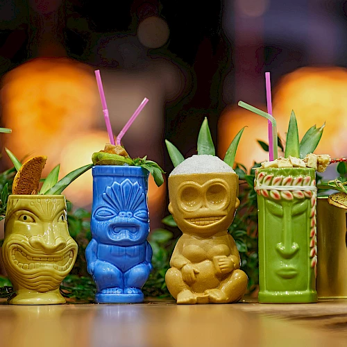 Colorful tiki mugs with straws are filled with tropical drinks, garnished with fruits and leaves, arranged on a table.