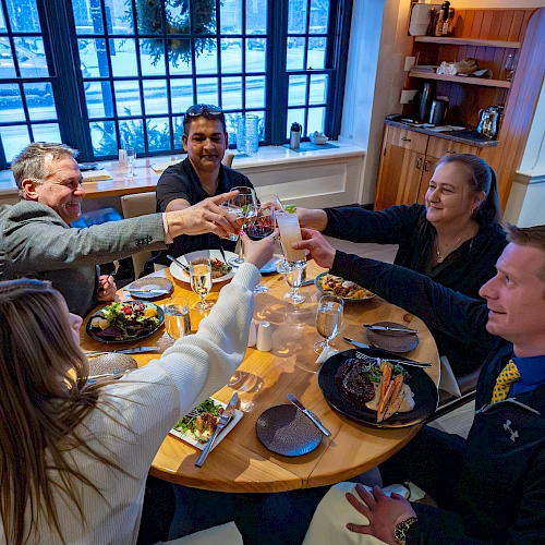 A group of five friends sits around a round table, toasting glasses, indoors at a cozy restaurant with large windows and plates of food.
