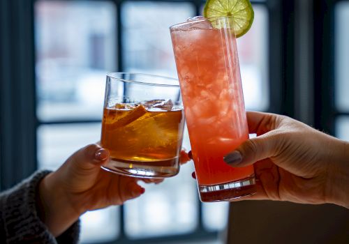 Two people toast with colorful cocktails: an amber drink and a tall pink-orange beverage with a lime wheel, clinking glasses by a window.
