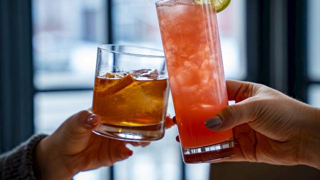 Two people toast with colorful cocktails: an amber drink and a tall pink-orange beverage with a lime wheel, clinking glasses by a window.