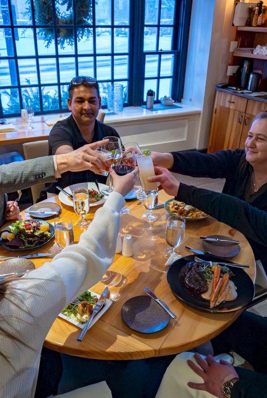A group of five friends around a round table toasting drinks in a bright, cozy restaurant with plates of food and large windows.
