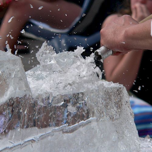 Ice blocks shattering into shards as hands smash through a blocky ice sculpture, splashes flying and onlookers watching intently.
