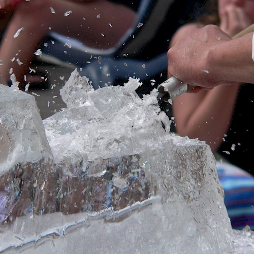 Ice blocks shattering into shards as hands smash through a blocky ice sculpture, splashes flying and onlookers watching intently.